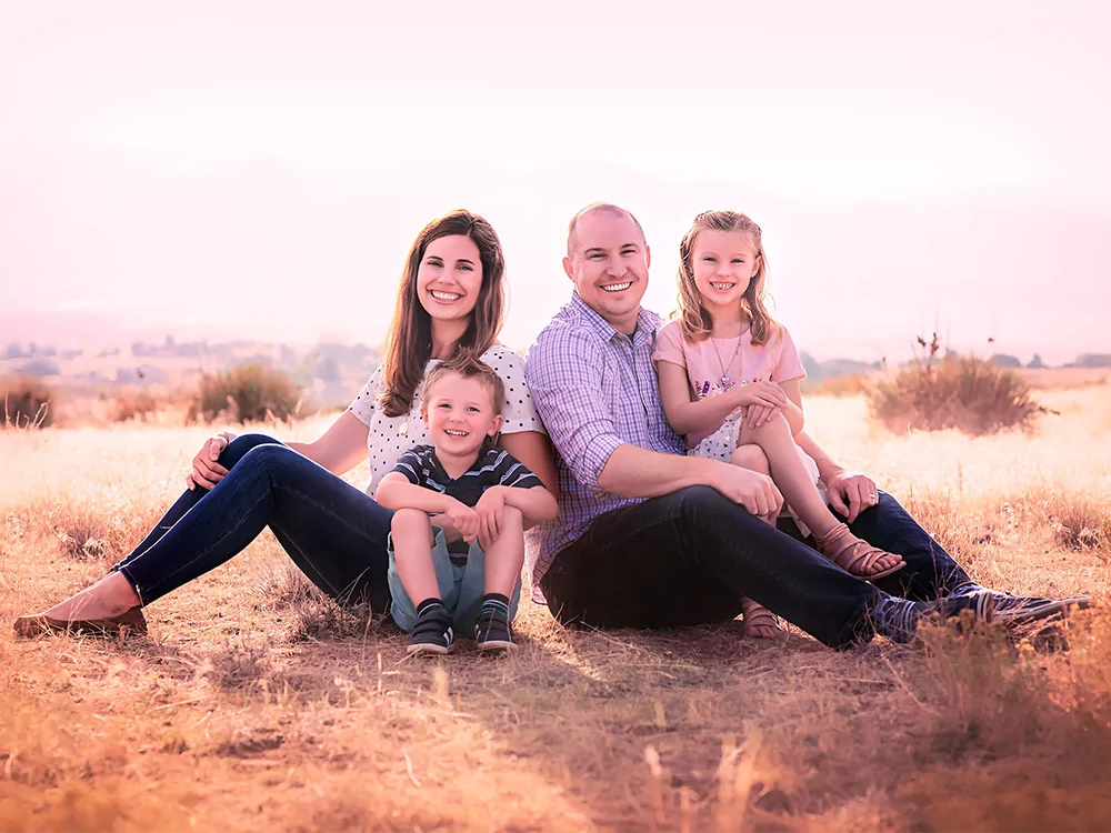 Family of four sits on dry grass in an open field during golden hour, with soft sunlight and distant hills in the background. The adults and children smile warmly, creating a serene and joyful outdoor scene.
