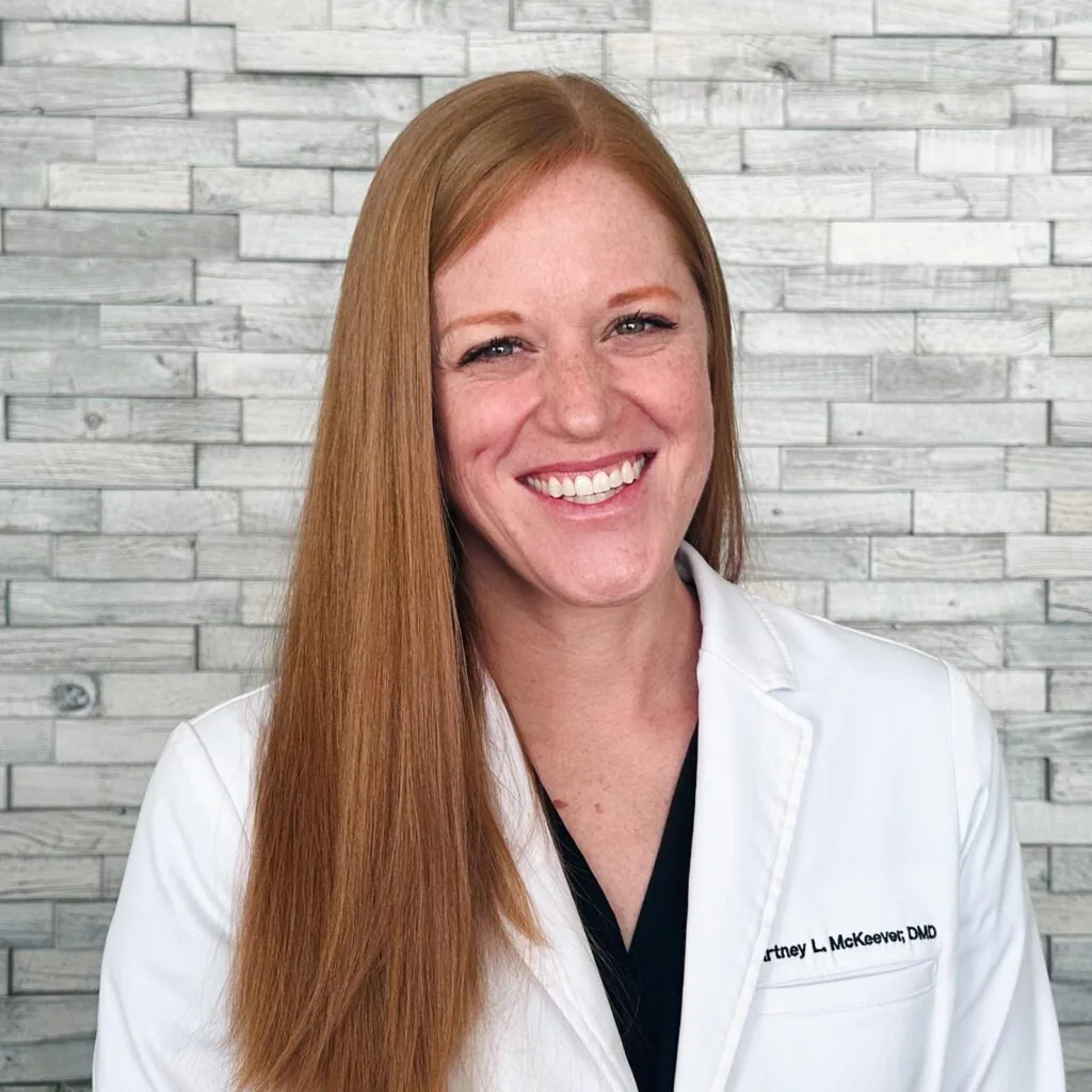 Portrait of Dr. Courtney McKeever with long, straight, reddish-brown hair wears a white lab coat embroidered with the name “Courtney L. McKeever, DMD.” They stand in front of a wall with horizontal light-colored bricks, suggesting a clinical or professional environment.