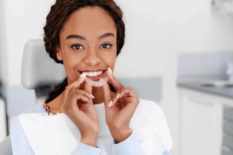 Woman seated in a dental chair holding an Invisalign near her mouth, wearing a dental bib in a modern clinic setting.