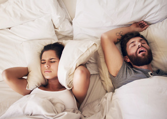Two people in bed, one covers ears with eyes shut while other sleeps on back with mouth open, white sheets and pillows.